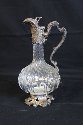 un pichet ancien en verre plaqué argent, posé sur un socle, sur fond noir