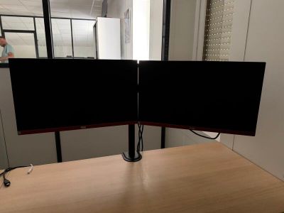 un bureau sur lequel sont posés deux écrans, avec un homme en arrière-plan
