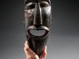 une personne tenant un masque en bois à la main, sur fond gris