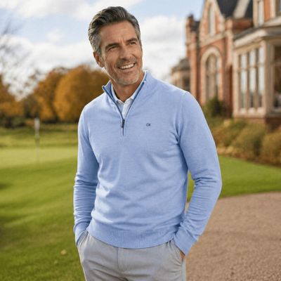 the man is standing in front of a large house wearing a blue sweater and grey pants