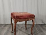 Un tabouret rembourré, dont l'assise est recouverte de tissu rouge, se trouve devant un rideau blanc.