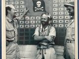 photographie en noir et blanc représentant trois hommes en uniforme militaire debout devant un mur orné d'un crâne et d'os croisés