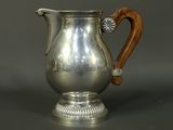 an antique silver pitcher with a wooden handle on a gray surface