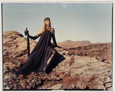 photo d'une femme vêtue d'une longue robe noire, assise sur un rocher, une épée à la main