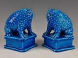 une paire de figurines de grenouilles en céramique bleue posées sur des socles, sur fond gris