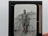 une main tenant une petite photo en noir et blanc représentant un homme devant une hutte au toit de chaume