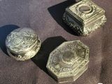 two small silver boxes sitting on a brown cloth
