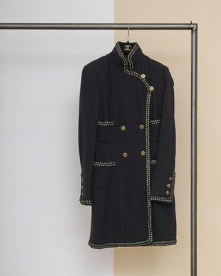 un manteau noir à boutons dorés suspendu à un portant devant un mur beige