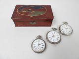 trois montres de poche en argent et un coffret en bois posés sur une surface blanche