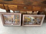 two framed paintings of a street scene on a wooden table