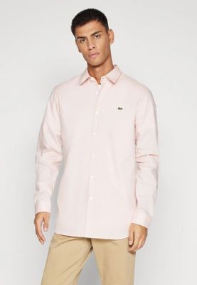 Un homme vêtu d'une chemise rose et d'un pantalon kaki se tient debout devant un mur blanc.
