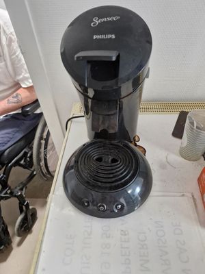 a coffee maker sitting on a table with a person in a wheel chair in the background