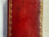 un vieux livre rouge avec des bordures dorées posé sur une surface blanche