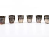 six verres à shot en argent alignés sur une surface blanche