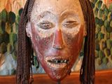 un masque en bois avec des dreadlocks, posé sur un socle devant un tableau