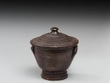 un vase marron muni d'un couvercle et d'une poignée, posé sur une surface blanche