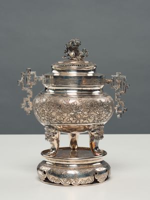 une théière ancienne en métal argenté, posée sur un support, sur une table blanche