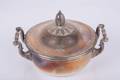 un bol ancien en métal argenté, muni d'un couvercle et de poignées, posé sur une surface blanche