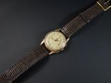 une image représentant une montre en or avec un bracelet en cuir marron sur un fond noir