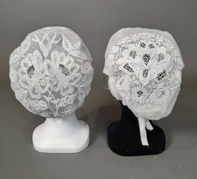 deux bonnets en dentelle blanche posés sur un support noir, sur fond gris