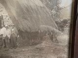 framed photograph of a group of people standing in front of a thatched house