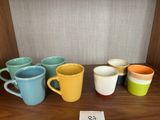 Plusieurs tasses à café de différentes couleurs sont alignées sur une étagère