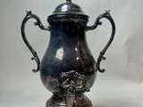 une cafetière ancienne en métal argenté, posée sur un socle et munie d'un couvercle