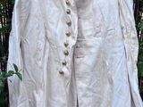 une veste beige à col et boutons rouges, suspendue à un cintre devant une clôture en bambou