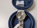 une montre en argent dans un étui bleu, sur lequel est accrochée une étiquette