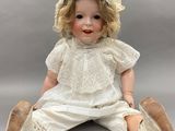 une poupée ancienne assise par terre, vêtue d'une robe blanche et d'un chapeau