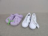 des chaussures Crocs violettes et blanches ornées de nœuds verts, posées sur un canapé