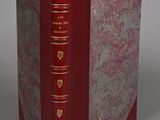 an old book with a red cover and gold trim sitting on a gray surface