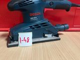 une ponceuse orbitale Bosch posée sur une table en bois, à côté d'une boîte rouge