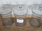 trois bocaux en verre transparent avec leurs couvercles sont posés sur une étagère en bois