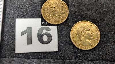 Trois pièces d'or sont posées sur un tissu noir, à côté d'une règle.