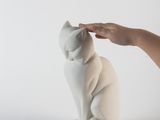 une statue de chat blanc posée sur une table blanche, à côté de la main d'une personne