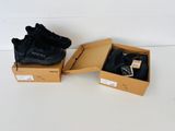 Deux paires de baskets noires se trouvent dans une boîte posée sur une surface blanche.