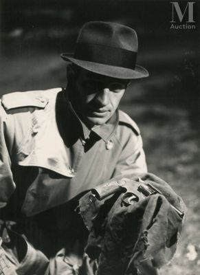 photo en noir et blanc d'un homme vêtu d'un trench-coat et d'un chapeau, tenant un gant de baseball