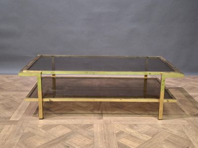Table basse en métal doré et verre fumé à deux plateaux. Tra…