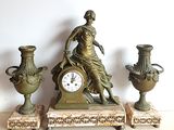 une statue représentant une femme assise sur une table, à côté de trois vases et d'une horloge