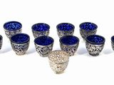 six blue glass cups and a silver vase on a white surface