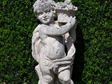 a statue of a boy holding a basket of flowers in front of a hedge