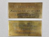 deux plaques en laiton posées sur un fond blanc, sur lesquelles figurent les noms de deux personnes