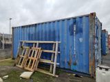 un conteneur d'expédition bleu posé à même le sol, à côté d'un tas de bois