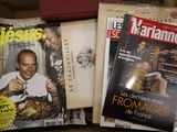 des magazines posés sur une table, devant lesquels se tient un homme tenant un couteau