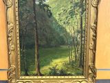 an oil painting of a forest scene in a gold frame on a blue surface