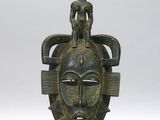 un gros plan d'un masque en bronze posé sur un socle, sur une surface blanche