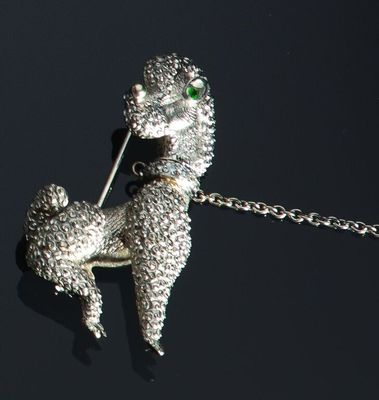 une image représentant une broche en forme de caniche argenté aux yeux verts, suspendue à une chaîne