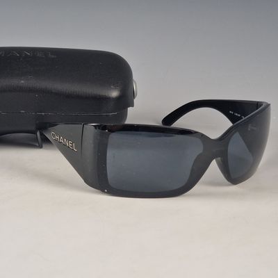 Lunettes de soleil Chanel à monture noire, avec leur étui, posées sur une surface blanche