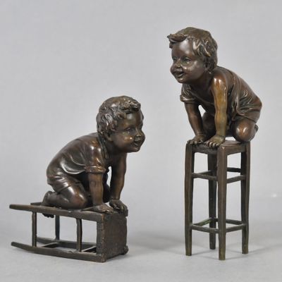 two bronze statues of children sitting on stools on a white surface